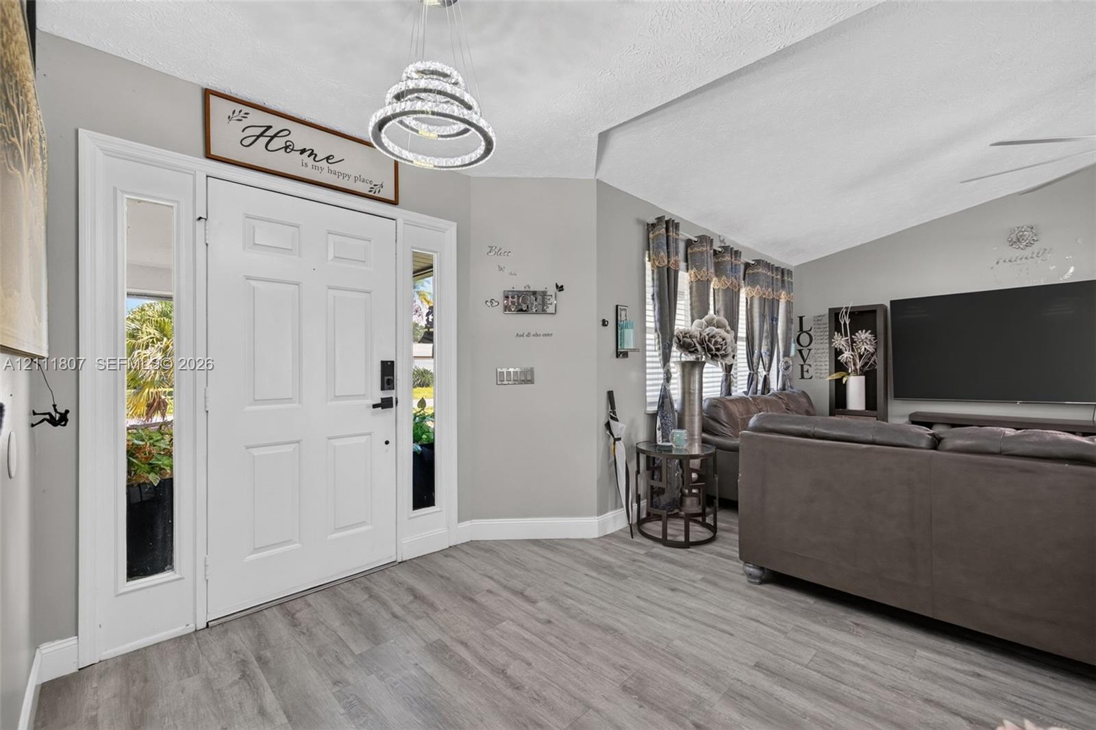 Entryway featuring a multi-paneled white door with dual sidelights, wood-finish flooring, vaulted ceiling, and a tiered crystal-style chandelier Entryway featuring a multi-paneled white door with dual sidelights, wood-finish flooring, vaulted ceiling, and a tiered crystal-style chandelier