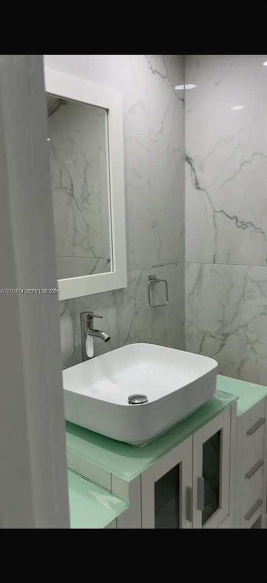 Bathroom featuring a vessel sink with a single-handle faucet, glass-top vanity with built-in cabinetry, framed mirror, and floor-to-ceiling veined tile wall surfacing