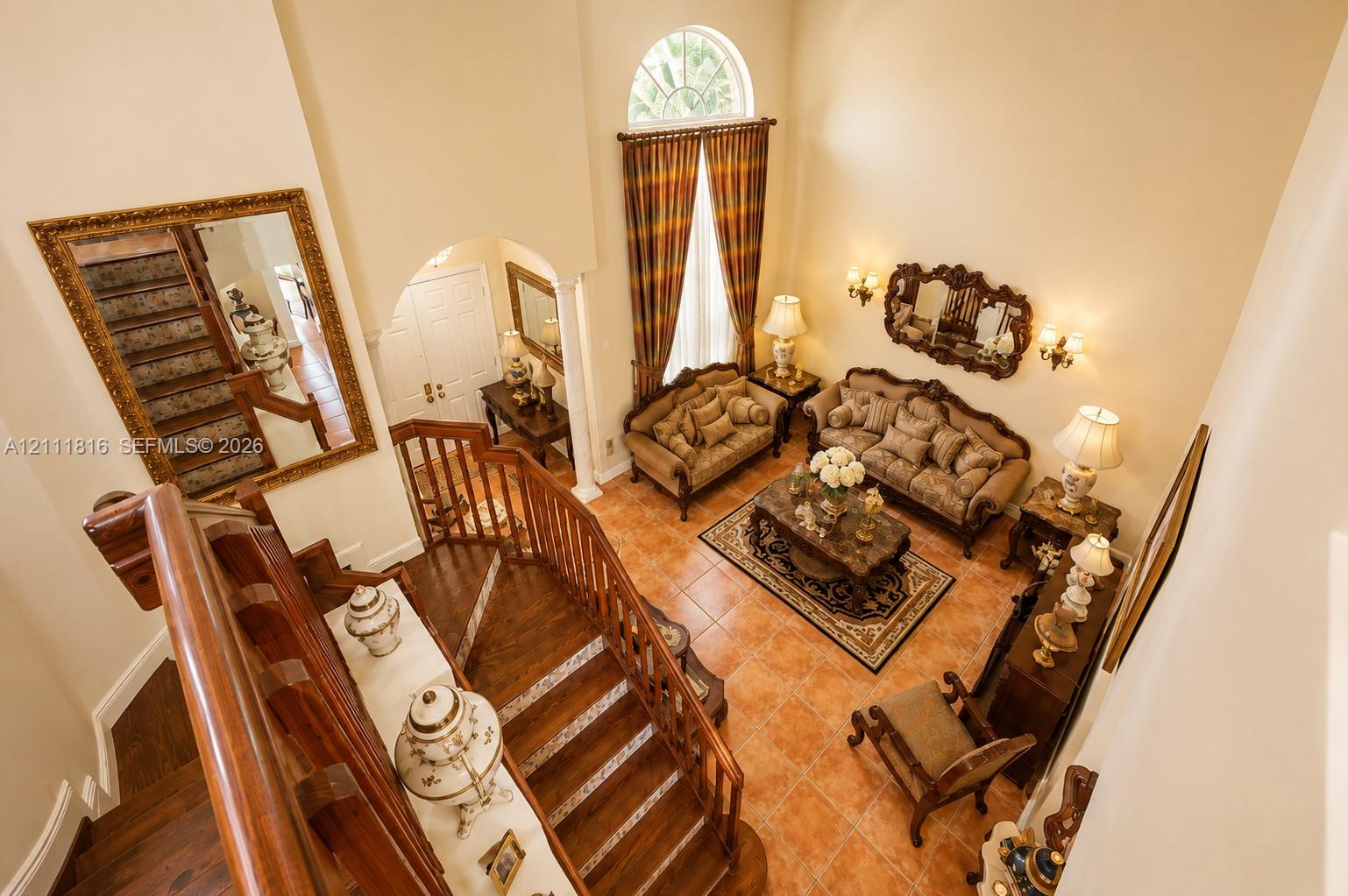 High-ceiling living area featuring tile flooring, a wood staircase with decorative balustrade, arched entryway, and an arched window