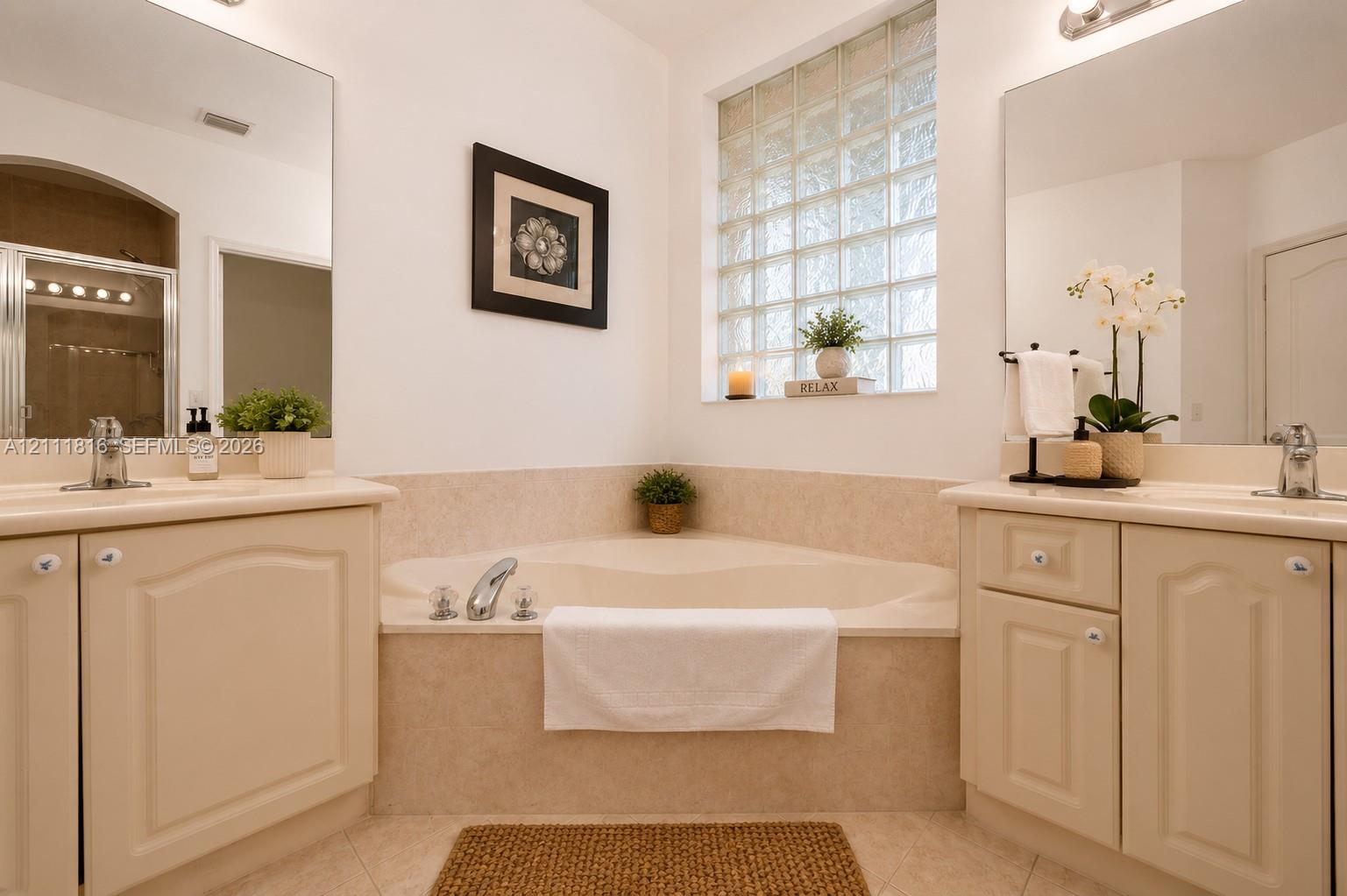 Dual vanities with cabinetry, built-in soaking tub, tile surround, and glass block window
