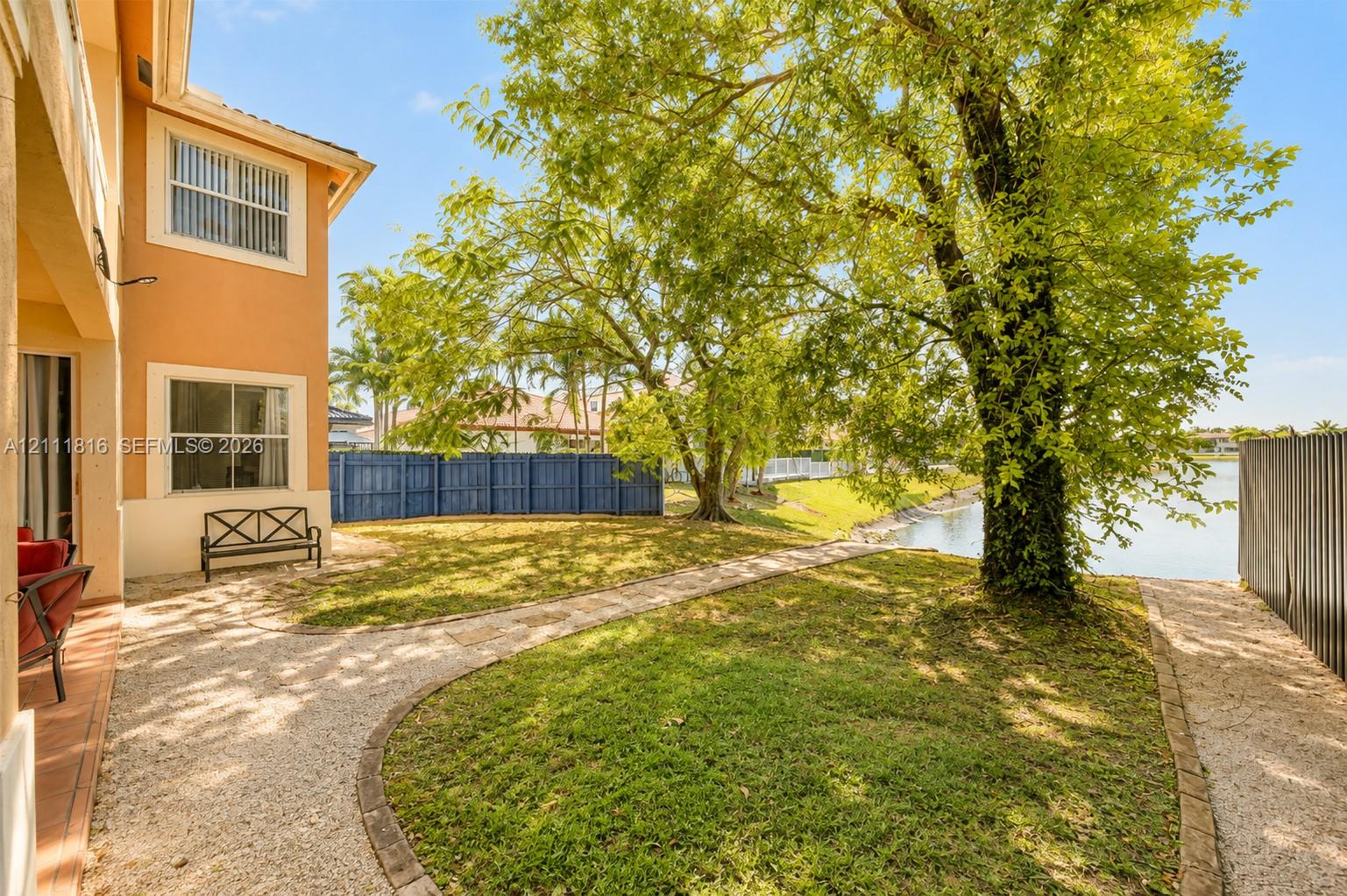 Waterfront backyard featuring a manicured lawn, stone-paved pathways, mature trees, and perimeter fencing