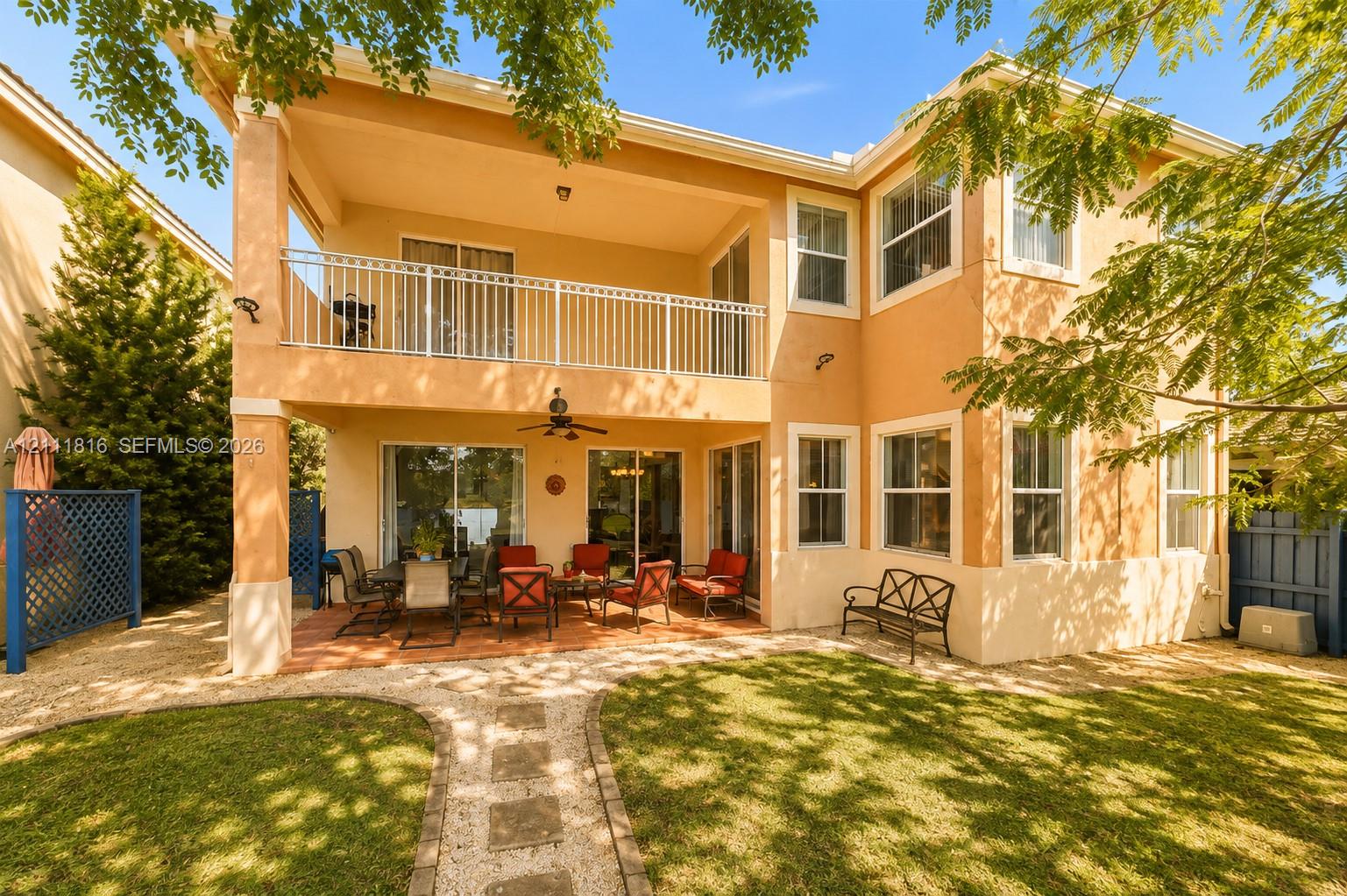 Two-story exterior with stucco finish, covered ground-level patio, second-story balcony with metal railing, stone-paver walkway, and manicured lawn