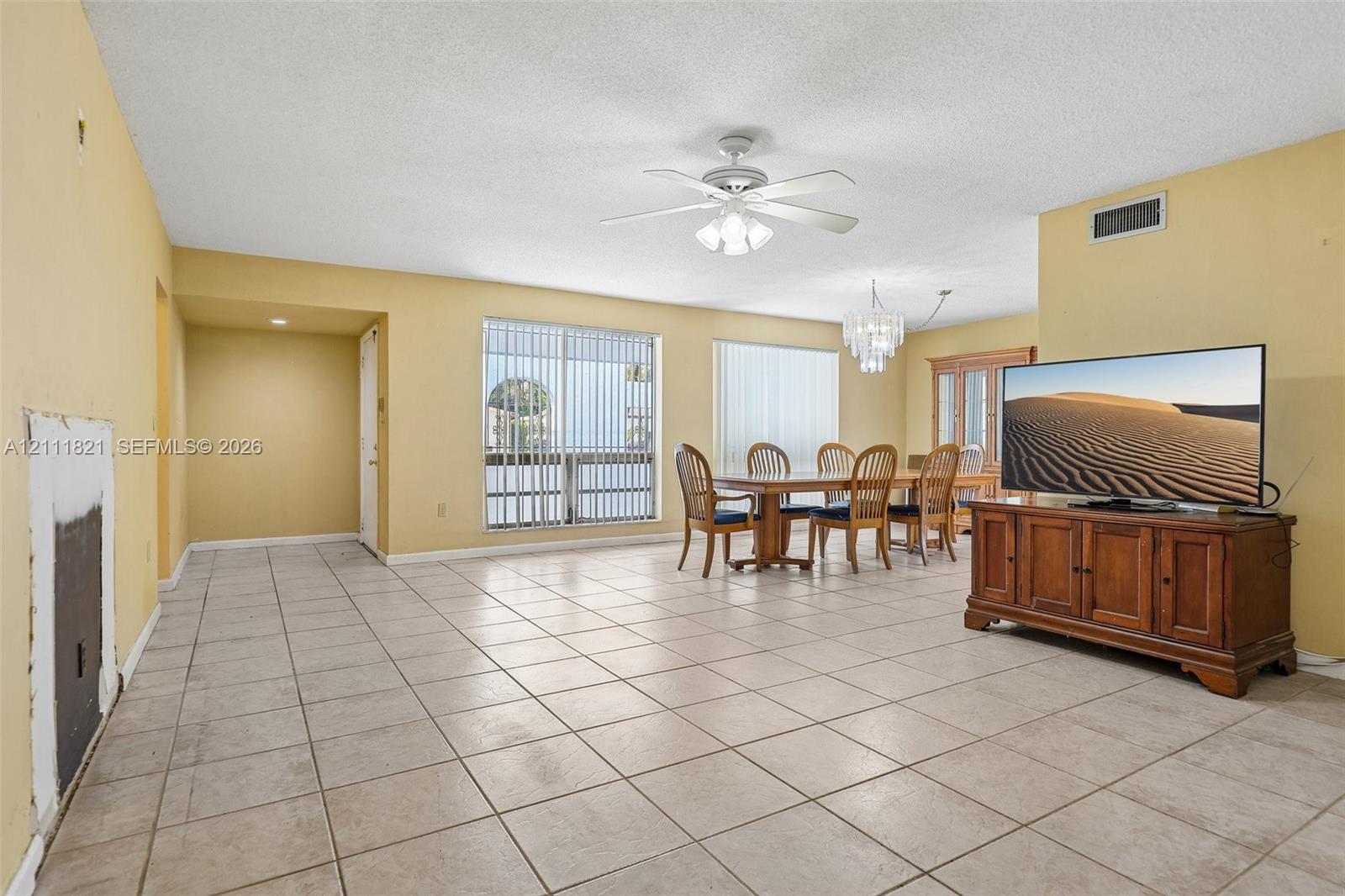 Neutral-toned tile flooring throughout the open-concept living area, featuring a decorative tiered chandelier, a white ceiling fan with integrated lighting, and large windows with vertical blinds