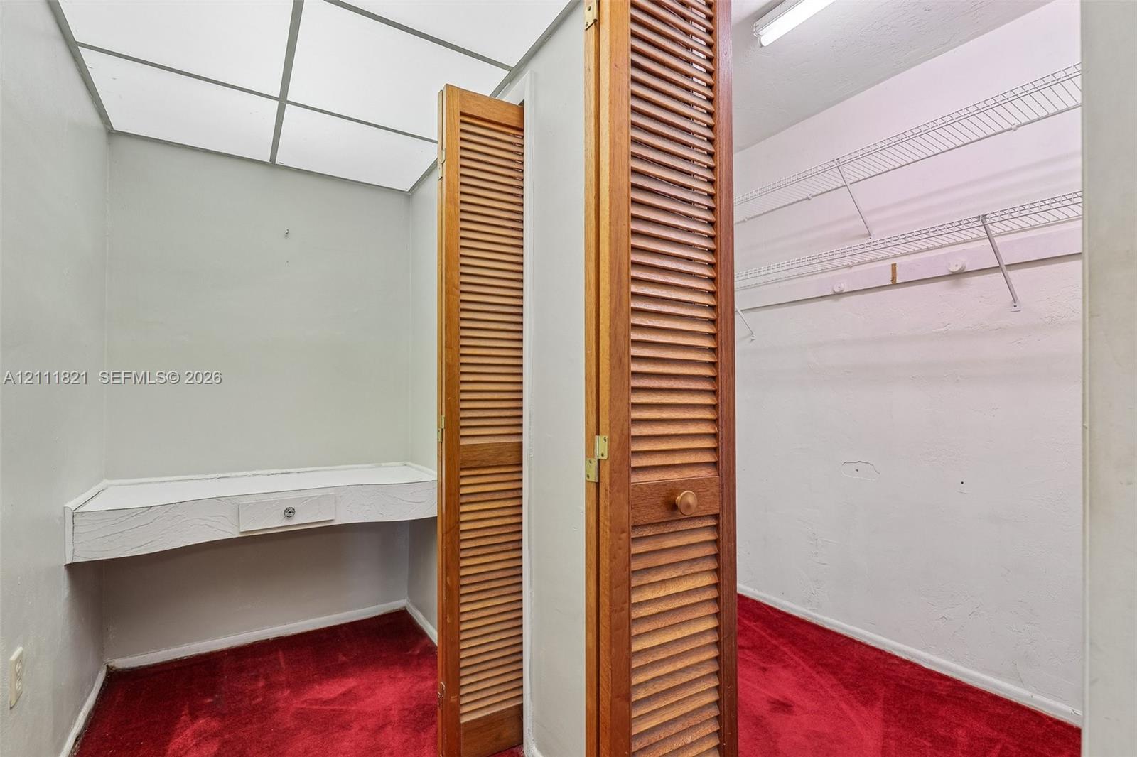 Storage space featuring louvered wooden bi-fold doors, white wall-mounted shelving, built-in vanity desk, and a grid-style ceiling light fixture