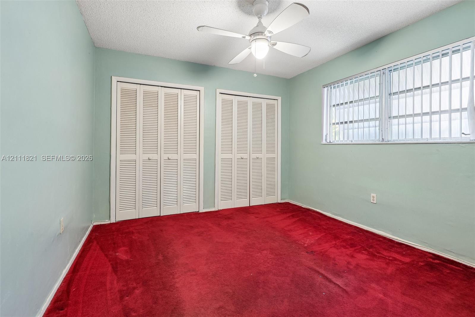 Room featuring carpeted flooring, dual closets with louvered bi-fold doors, a multi-pane horizontal window, and a central ceiling fan