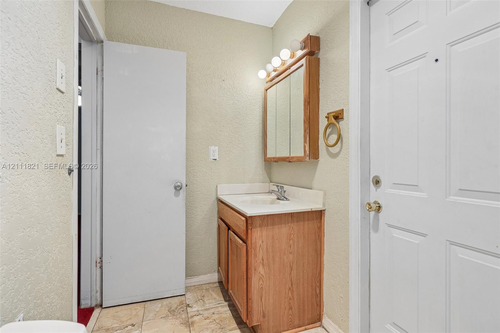 Bathroom vanity featuring a wooden cabinet base, integrated sink with faucet, surface-mounted medicine cabinet, four-bulb vanity light fixture, and stone-finish floor tiling
