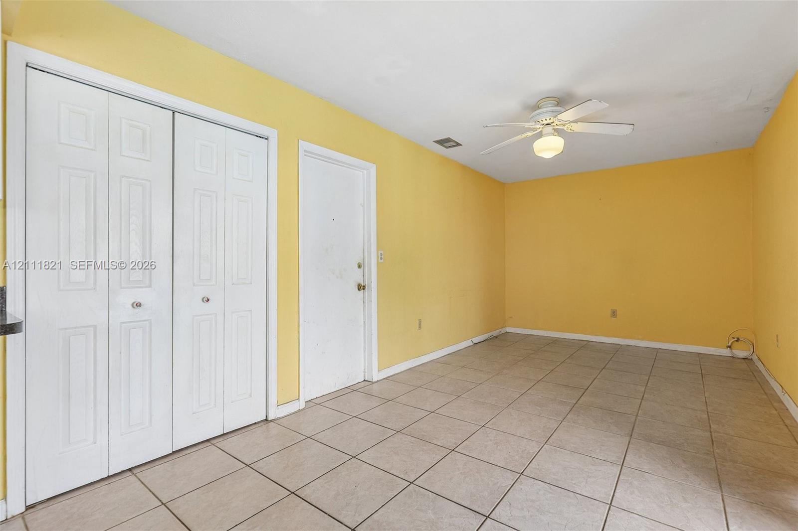 Interior space featuring neutral-tone tile flooring, white bi-fold closet doors, a single white swing door, and a ceiling-mounted light fixture with fan blades