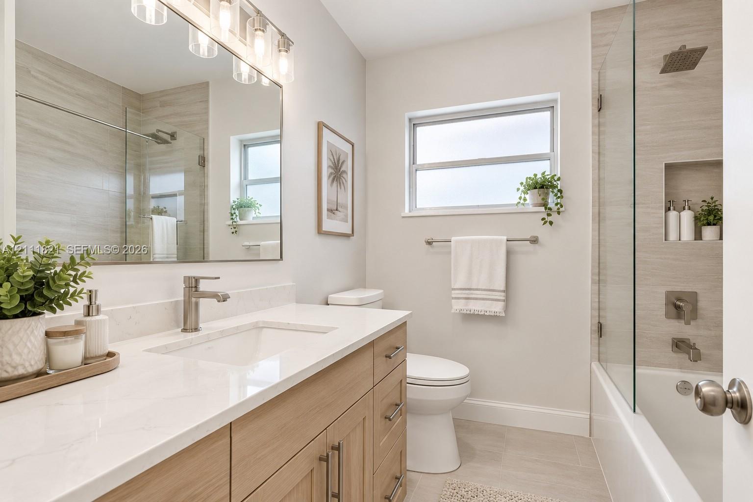 Bathroom featuring a vanity with wood-finish cabinetry, white countertop with integrated sink, rectangular mirror with multi-light fixture, and a glass-enclosed shower-tub combination with horizontal tile surround and built-in recessed shelf