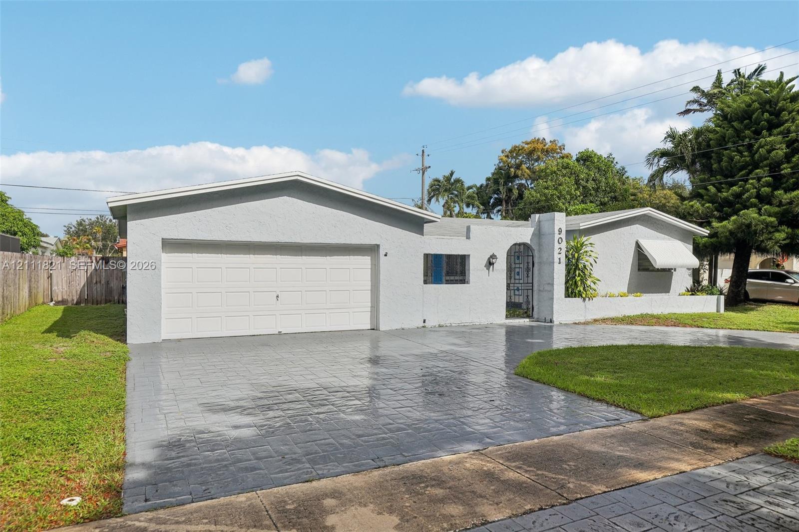 Single-story exterior featuring a light-toned stucco facade, dual-car garage, stamped concrete driveway, metal entryway gate, and awning-style window covers