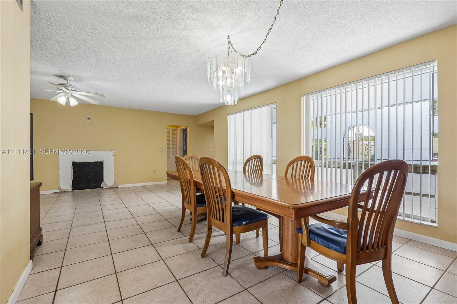 Open-concept living space featuring ceramic tile flooring, a built-in masonry fireplace, a tiered crystal chandelier, and large windows with vertical blinds