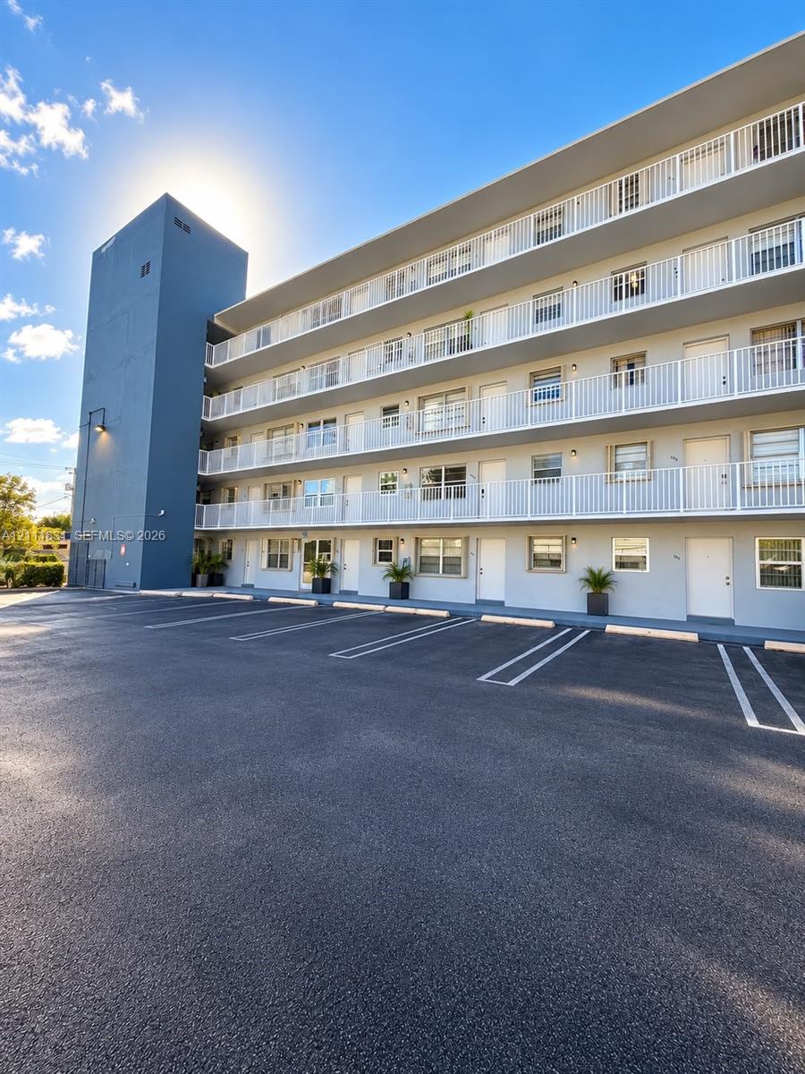 Multi-story exterior featuring a smooth stucco finish, asphalt-paved parking area, white metal balcony railings, and individual unit entry doors