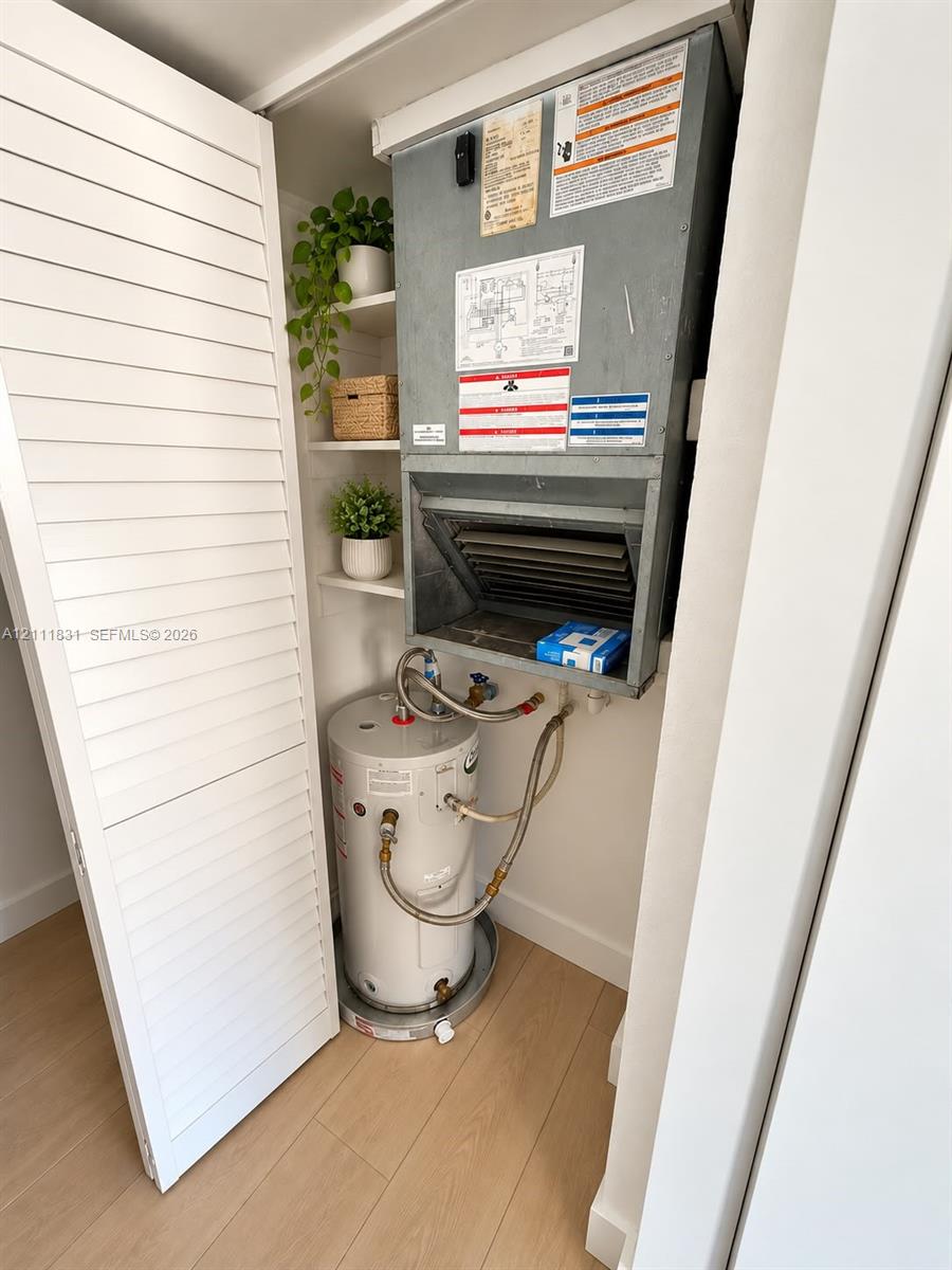 Utility closet featuring an electric water heater, wall-mounted air handling unit, and built-in shelving behind louvered doors with wood-finish flooring