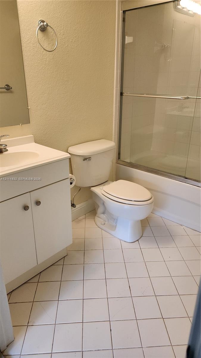 Bathroom featuring a vanity with an integrated sink, a porcelain toilet, a tub-and-shower combination with a sliding glass enclosure, and tiled flooring