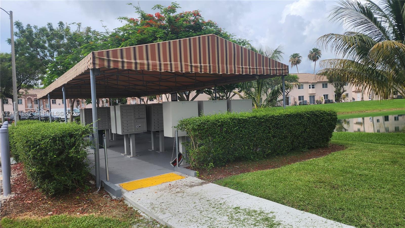 Covered centralized mail delivery station with metal pedestal-mounted compartments, situated on a concrete pad with a striped fabric canopy