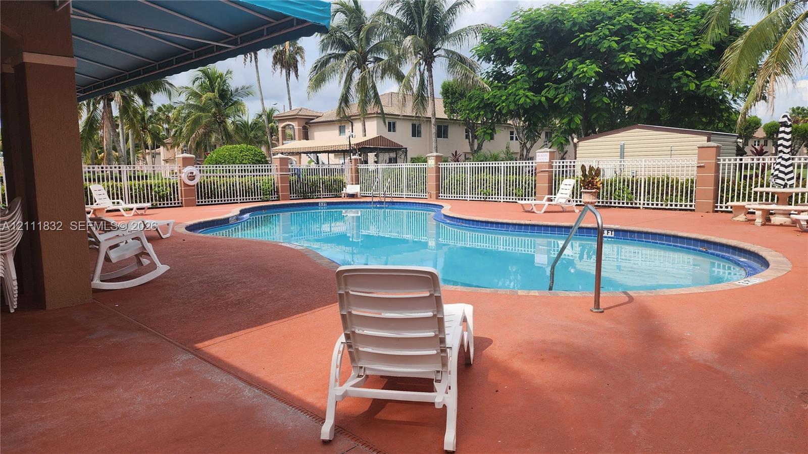 In-ground swimming pool with tile border, paver-style decking, white metal perimeter fencing, and retractable fabric awning