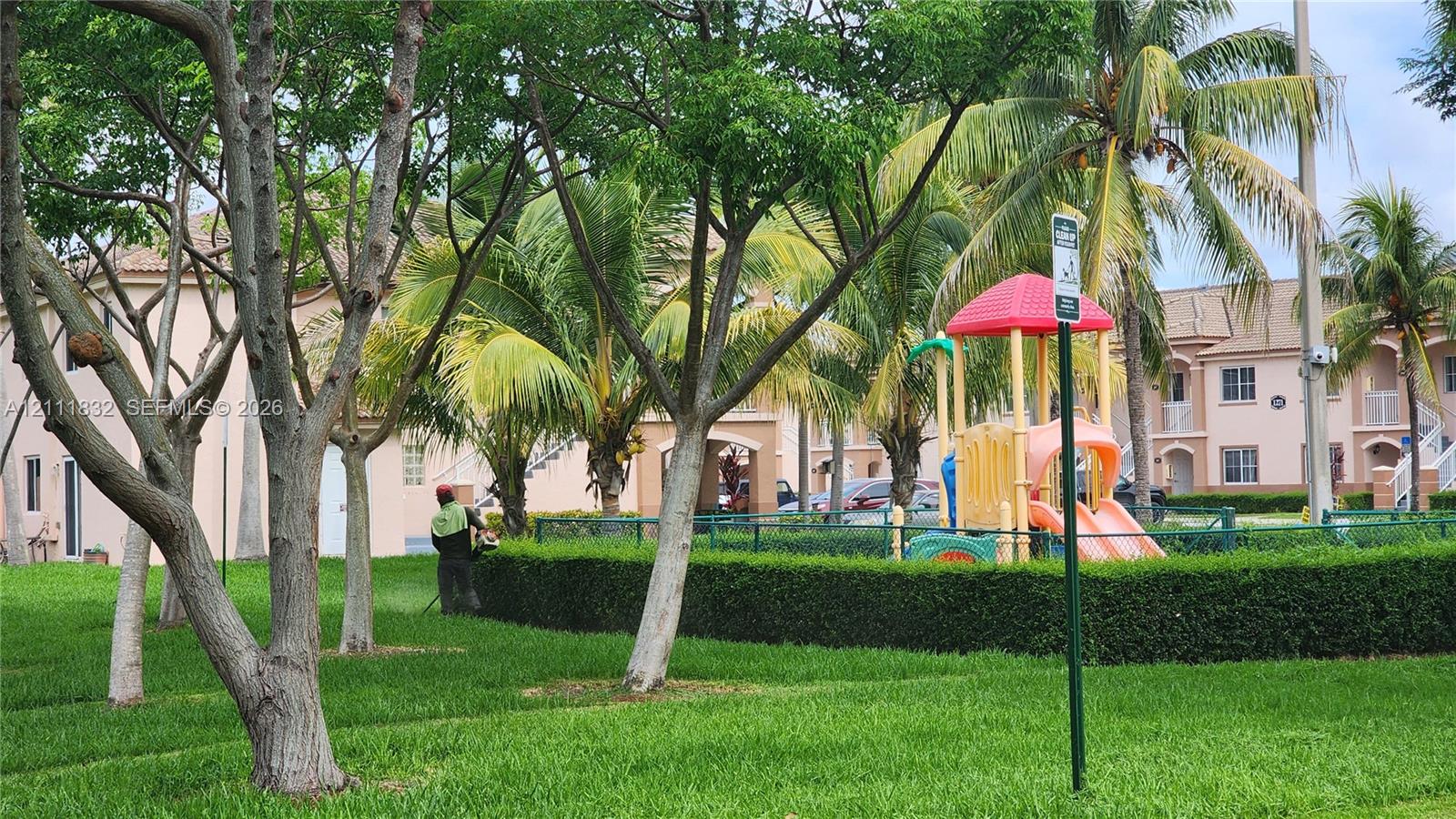 Communal green space featuring manicured lawns, mature tropical landscaping, and a fenced playground structure