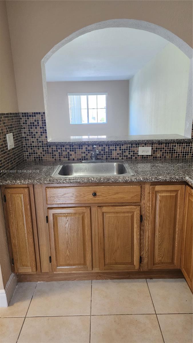 Kitchen pass-through featuring an arched opening, stone-finish countertops, stainless steel sink, mosaic tile backsplash, and wood-finish cabinetry