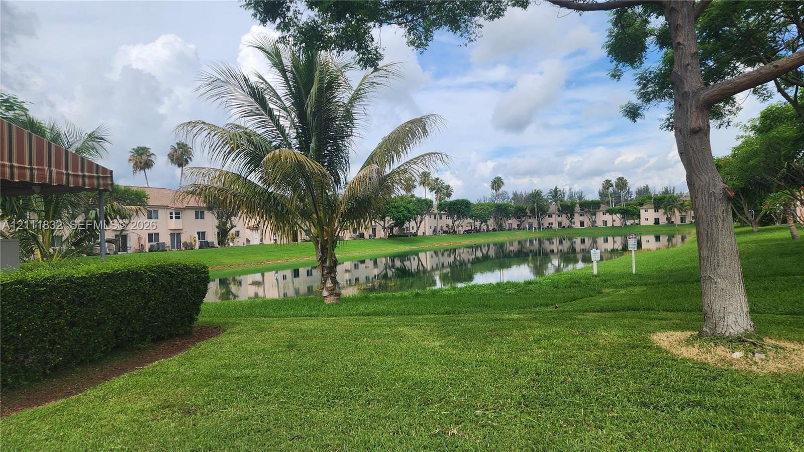 Waterfront grounds featuring a manicured lawn, established palm trees, mature canopy trees, and a central pond