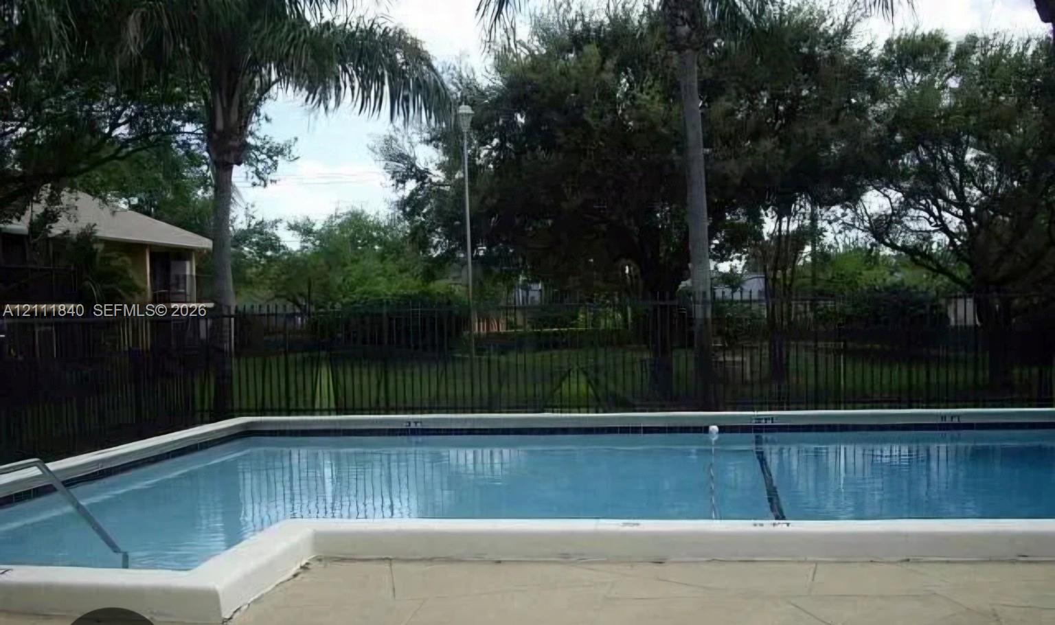 Swimming pool with a concrete pool deck, metal perimeter fencing, and mature trees