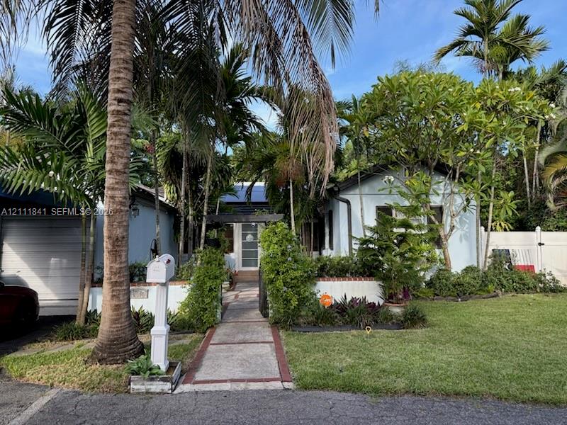 Light-colored exterior siding with a covered entryway, paved walkway, brick-accented landscape border, and mature tropical landscaping