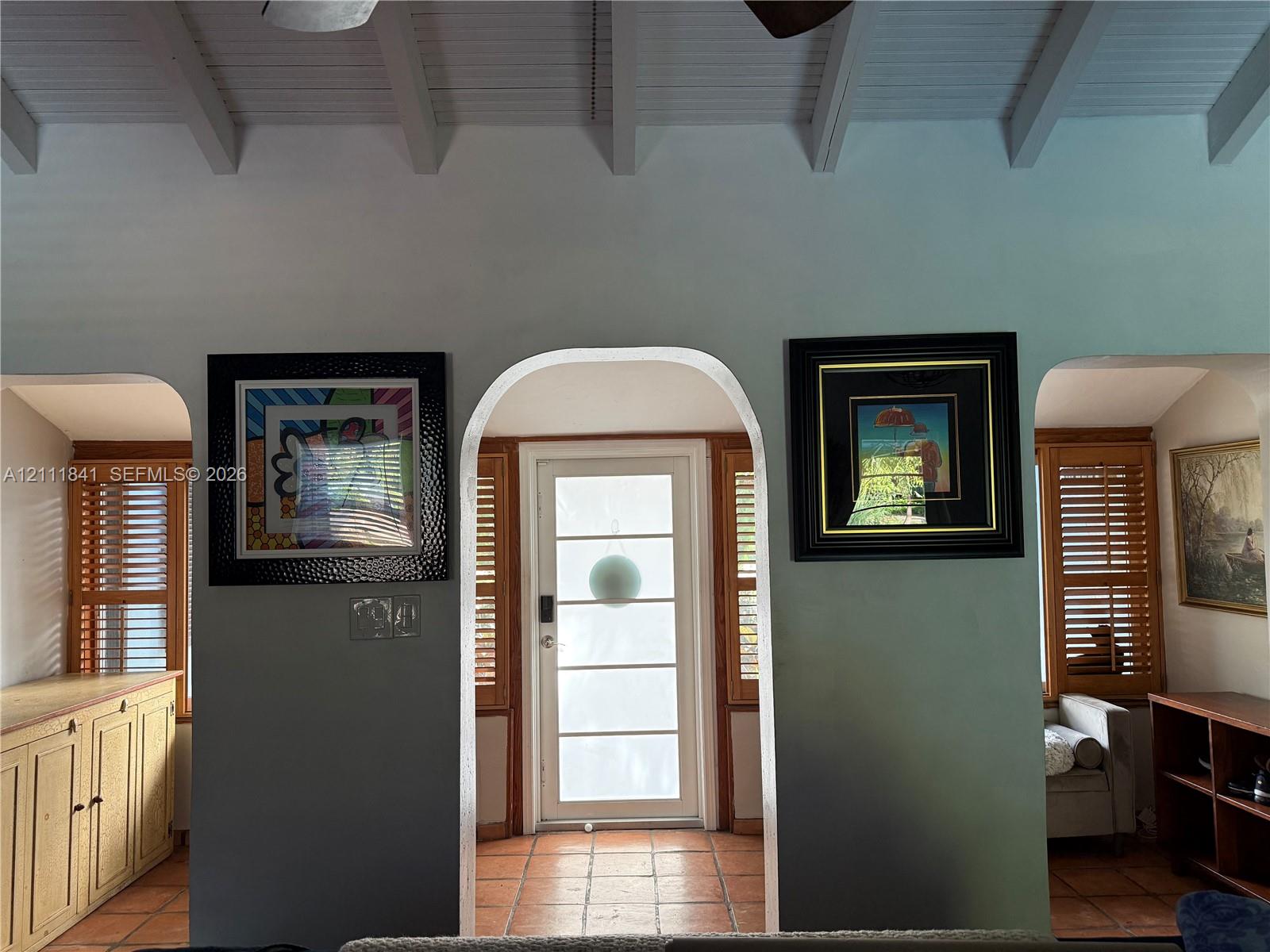 Exposed beamed ceiling with white finish, terracotta tile flooring, arched doorways, and natural wood louvered shutters