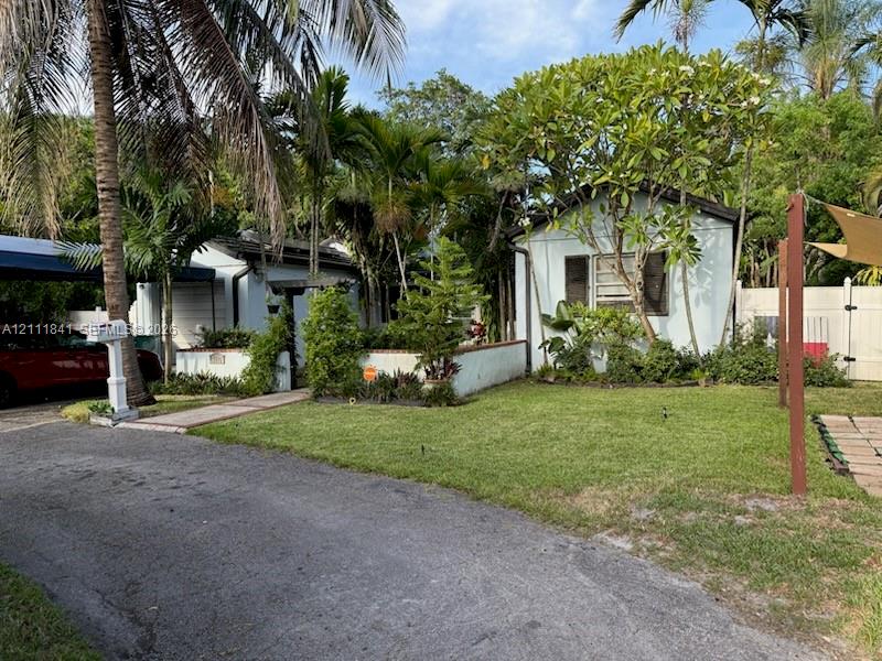 Light-colored exterior siding with shuttered windows, complemented by a front yard with mature landscaping, a paved walkway, a metal-framed carport, and perimeter privacy fencing