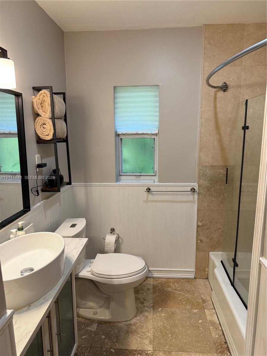 Bathroom featuring a vessel sink with a stone-top vanity, tile flooring, glass shower panel with a curved curtain rod, and wainscoting