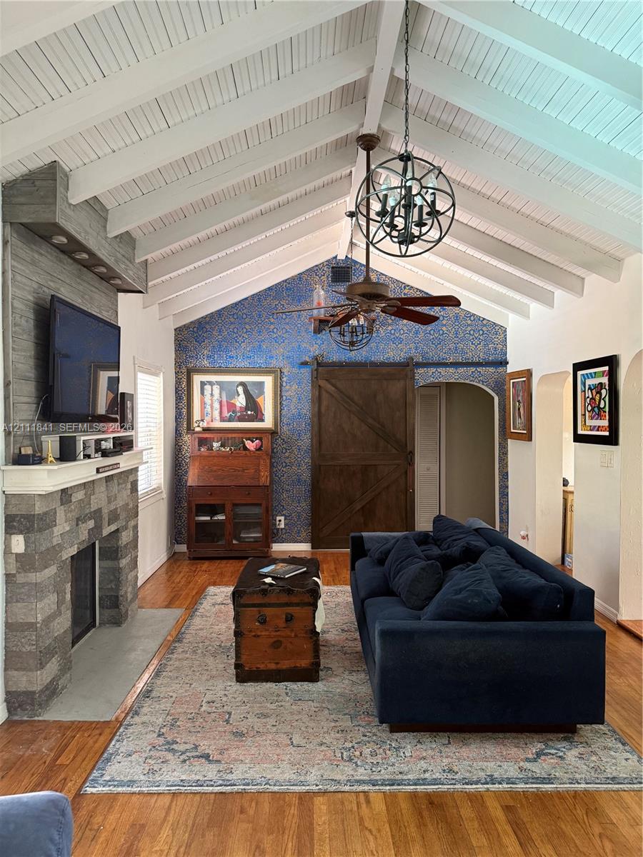 Vaulted ceiling with exposed white beams, stone-surround fireplace, wood-finish flooring, and sliding wood barn door