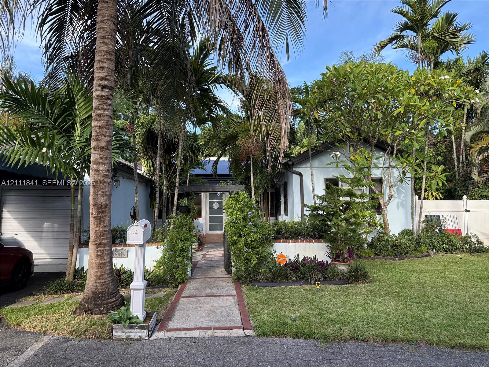 Single-story exterior featuring a paved walkway, attached garage, white privacy fencing, and lush mature landscaping