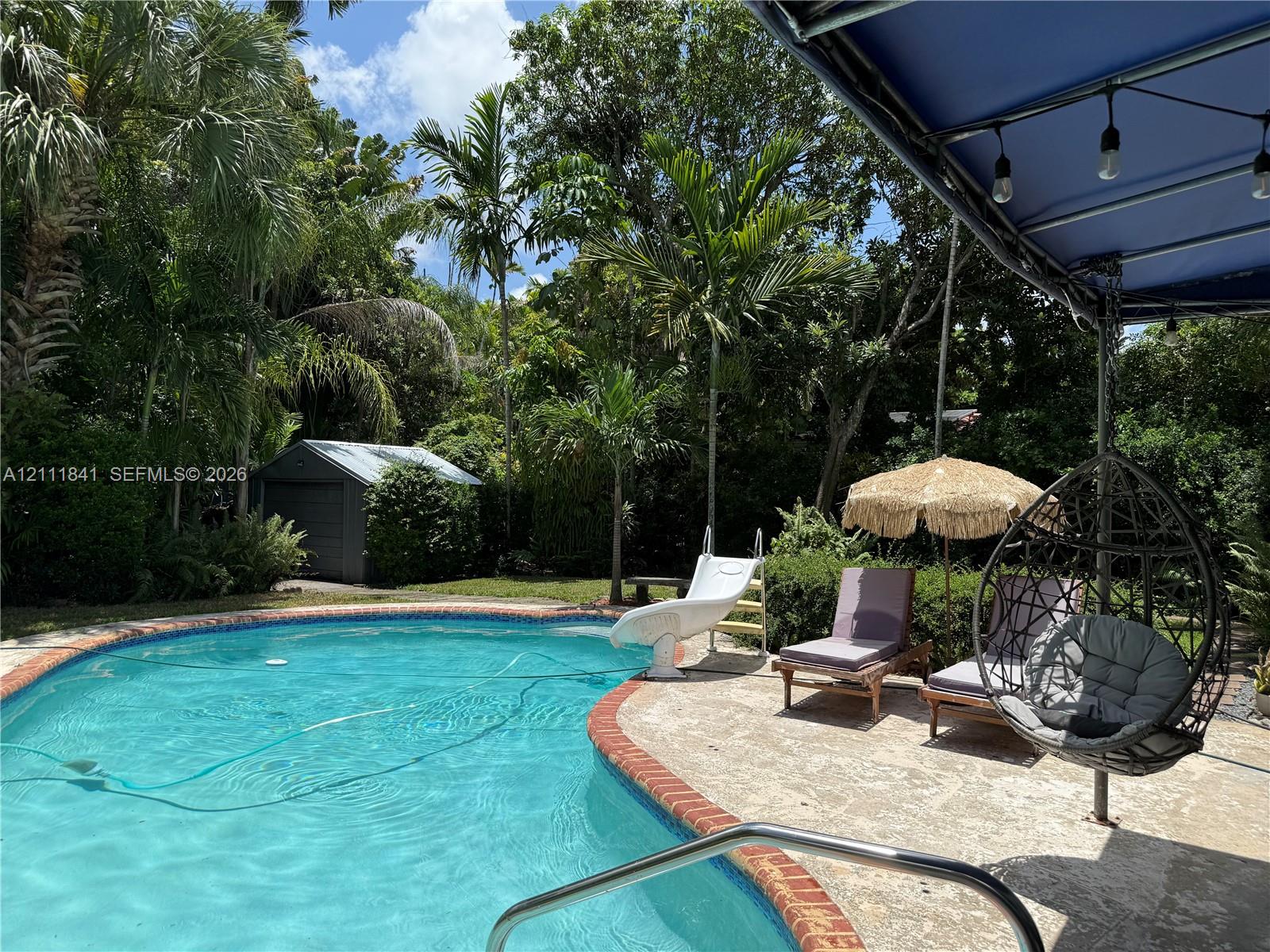 In-ground swimming pool with a red brick coping, concrete pool deck, attached pool slide, and a metal storage shed