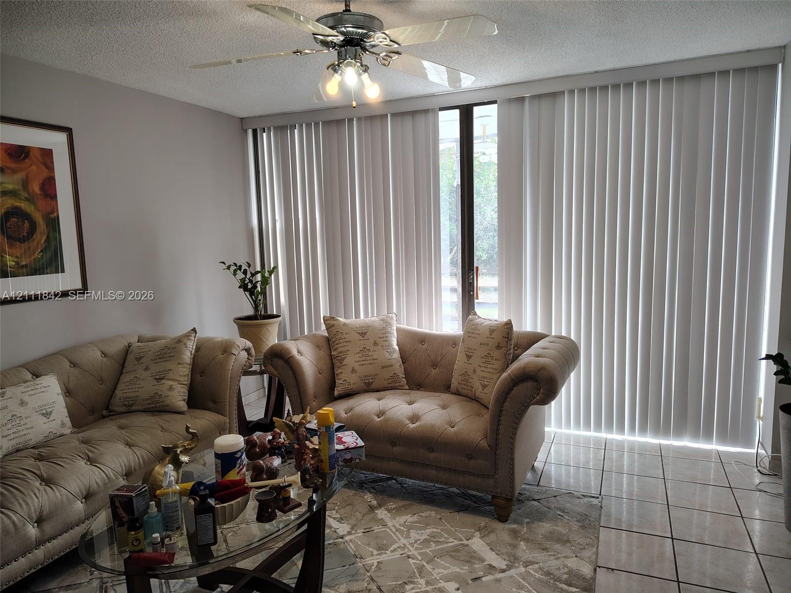 Living area featuring large-format ceramic tile flooring, floor-to-ceiling vertical blinds, and a central ceiling fan with integrated lighting