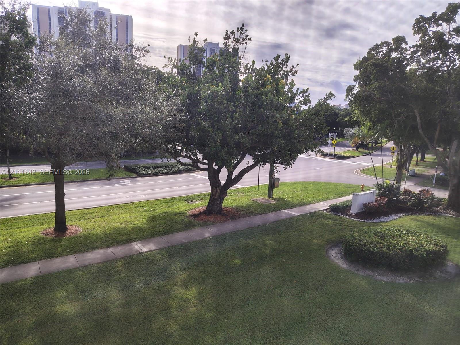 Expansive green space featuring mature trees, manicured lawn, concrete sidewalk, and landscaped garden beds