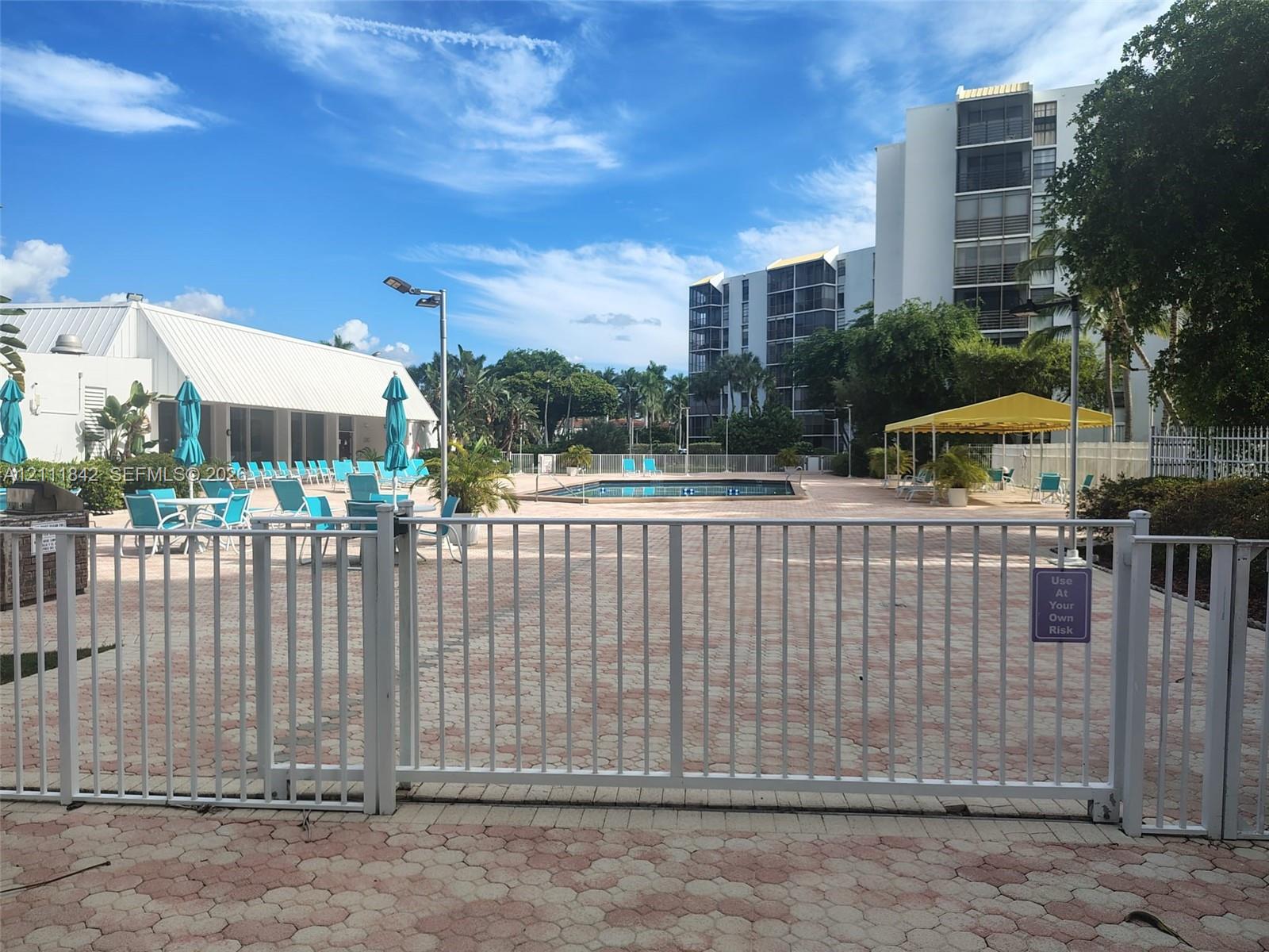 Community pool area featuring paver decking, white metal security fencing, and a yellow-canopied pavilion