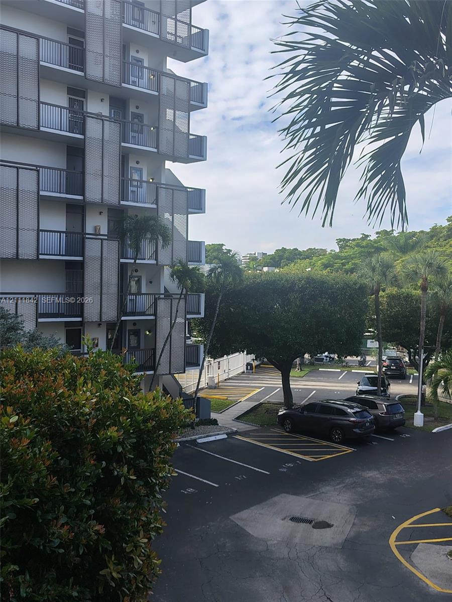 Multi-story building exterior featuring staggered balconies with metal mesh privacy screening, dedicated asphalt parking area, and mature palm landscaping