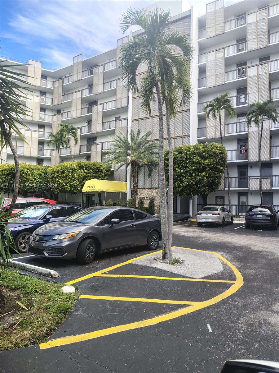 Multi-story residential building featuring a concrete facade with decorative metal screening, private balconies, paved surface parking, and mature palm landscaping