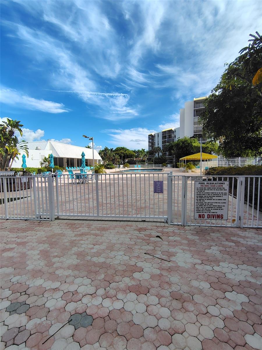 Community swimming pool area featuring paved decking, metal perimeter fencing, and a covered pavilion