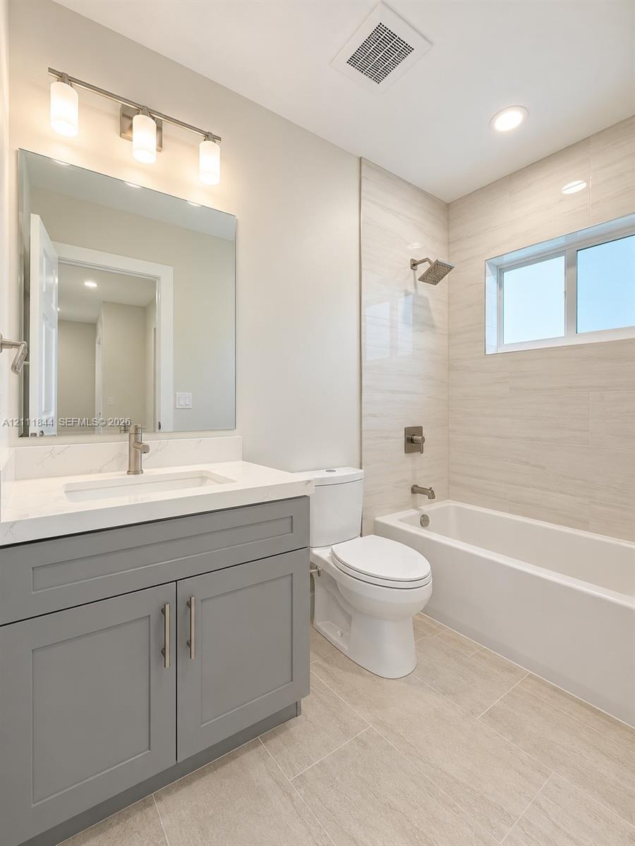 Bathroom featuring a grey shaker-style vanity with a white countertop, large frameless mirror, three-light vanity fixture, and a tile-surround tub with metallic plumbing fixtures
