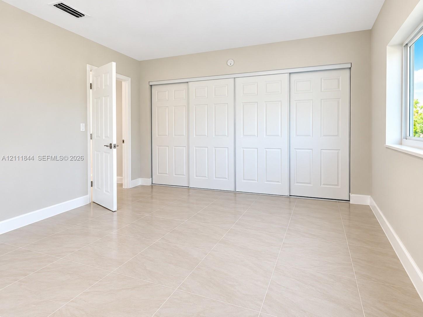 Neutral-toned room featuring large-format tile flooring, oversized four-panel sliding closet doors, and a single window