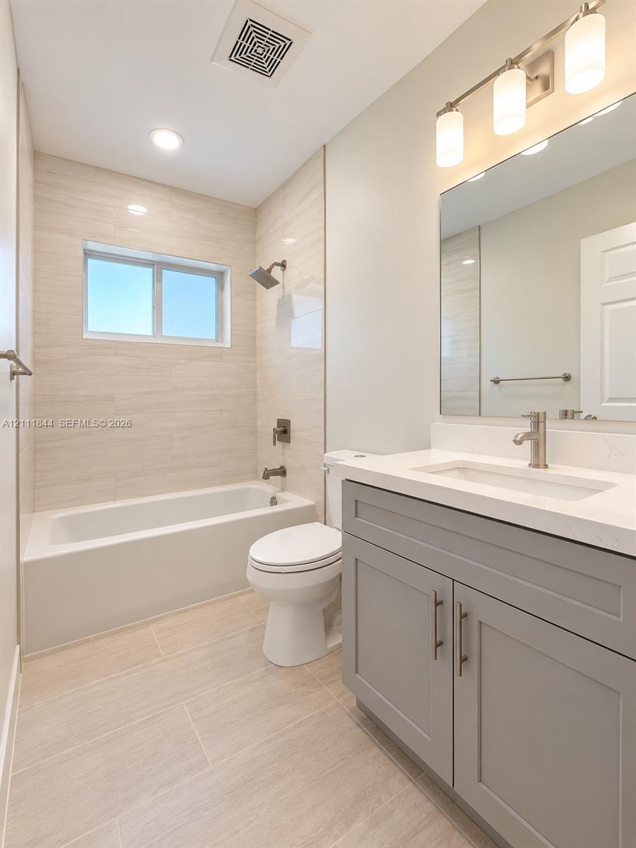 Bathroom featuring a gray vanity with a stone countertop, an integrated rectangular sink, neutral large-format wall and floor tile, and a bathtub with showerhead and mixer faucet