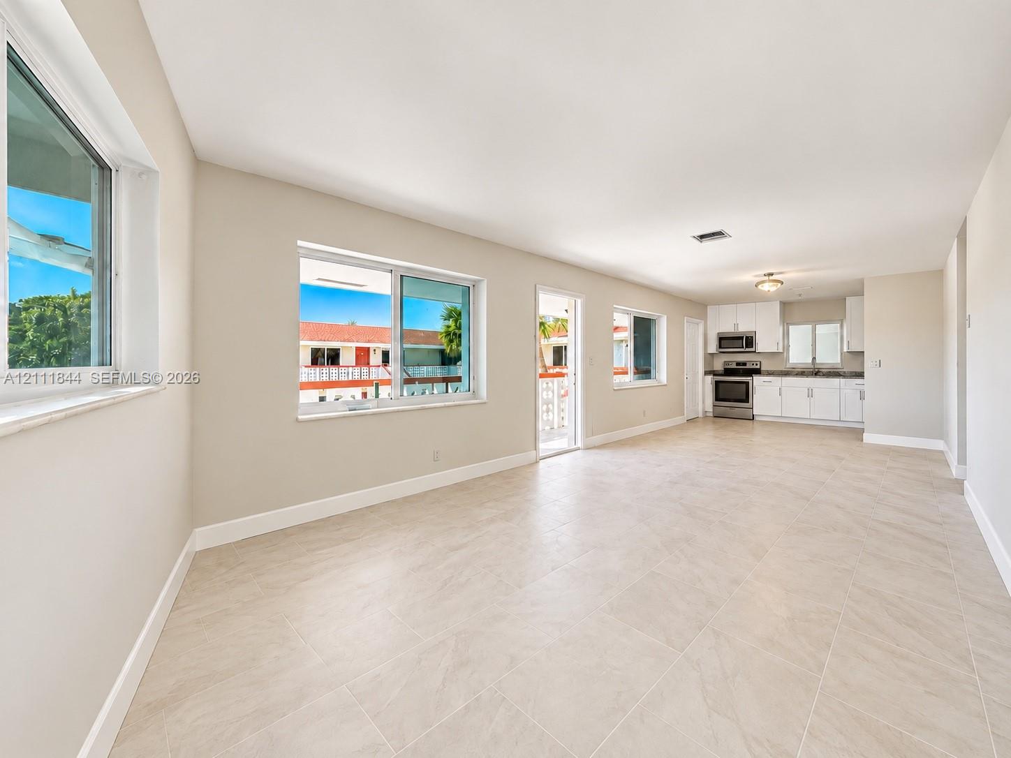 Open-concept floor plan featuring neutral large-format tile flooring, multiple windows, white kitchen cabinetry, and stainless steel appliances
