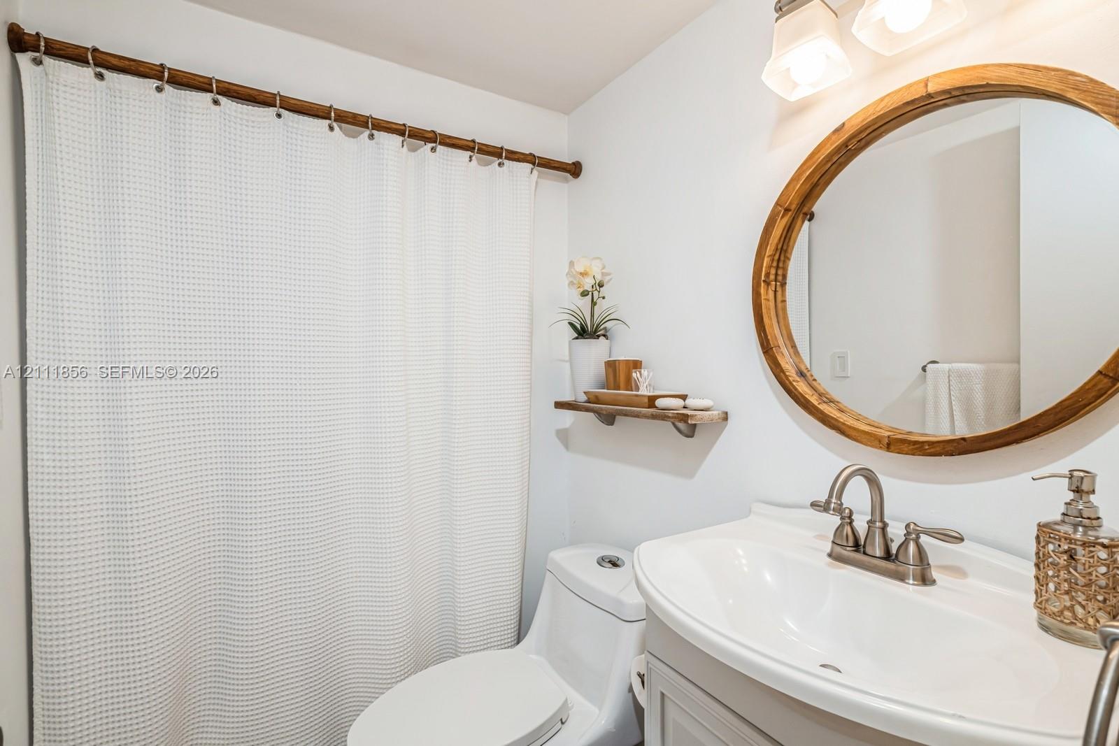 Bathroom vanity with a white basin and metallic faucet, circular wood-framed mirror, dual-bulb wall-mounted light fixture, and wooden wall-mounted shelving