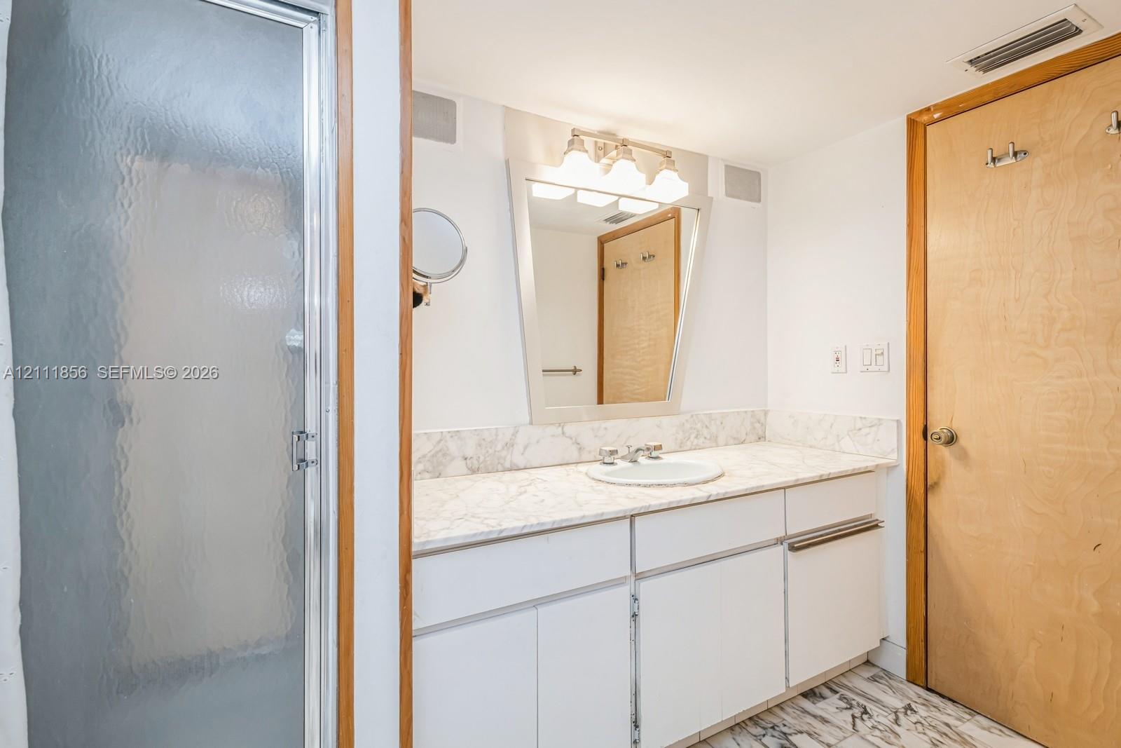 Bathroom featuring a vanity with stone-finish countertop, integrated sink, framed wall mirror with multi-bulb overhead lighting, stone-patterned flooring, and a glass-enclosed shower