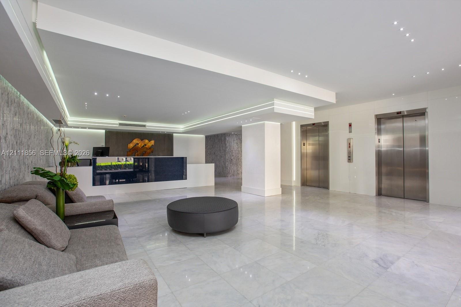 Lobby area featuring polished stone flooring, recessed ceiling lighting, cove accent lighting, stainless steel elevator doors, and a built-in reception desk