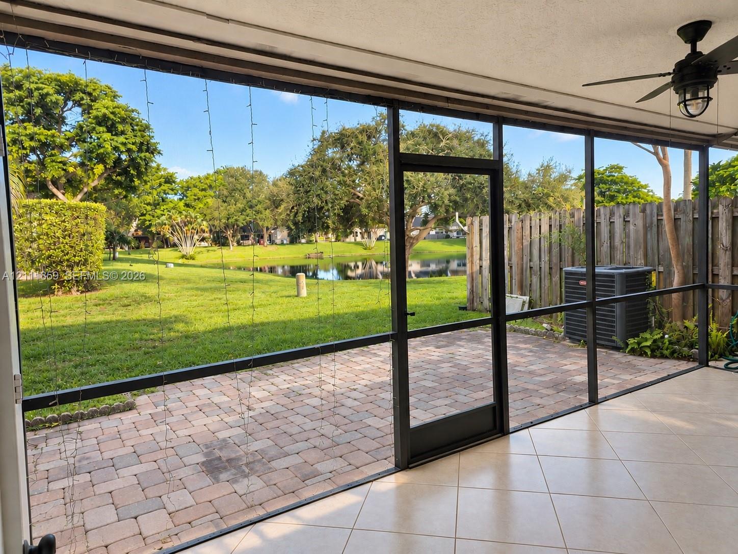 Screened-in patio with tiled flooring and ceiling-mounted fan, featuring brick-paver hardscaping, mature landscaping, and waterfront scenery