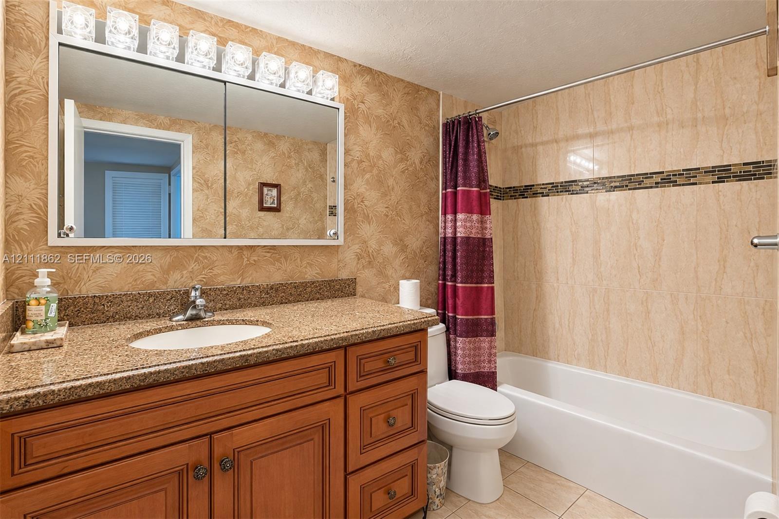 Bathroom featuring wood-finish cabinetry, stone-texture countertop, integrated vanity sink, multi-light fixture, and tub-shower combination with decorative tile inlay