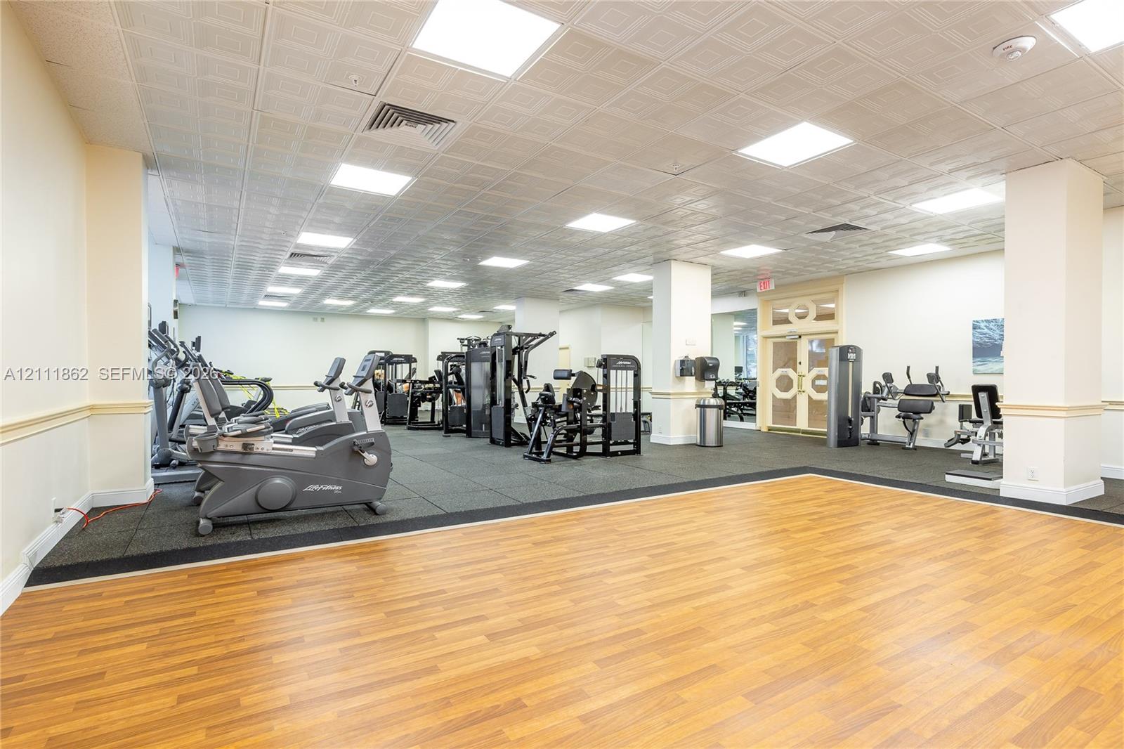 Spacious fitness center featuring wood-finish flooring, a rubberized exercise surface, recessed ceiling lighting, a decorative coffered ceiling, and glass-paneled double doors