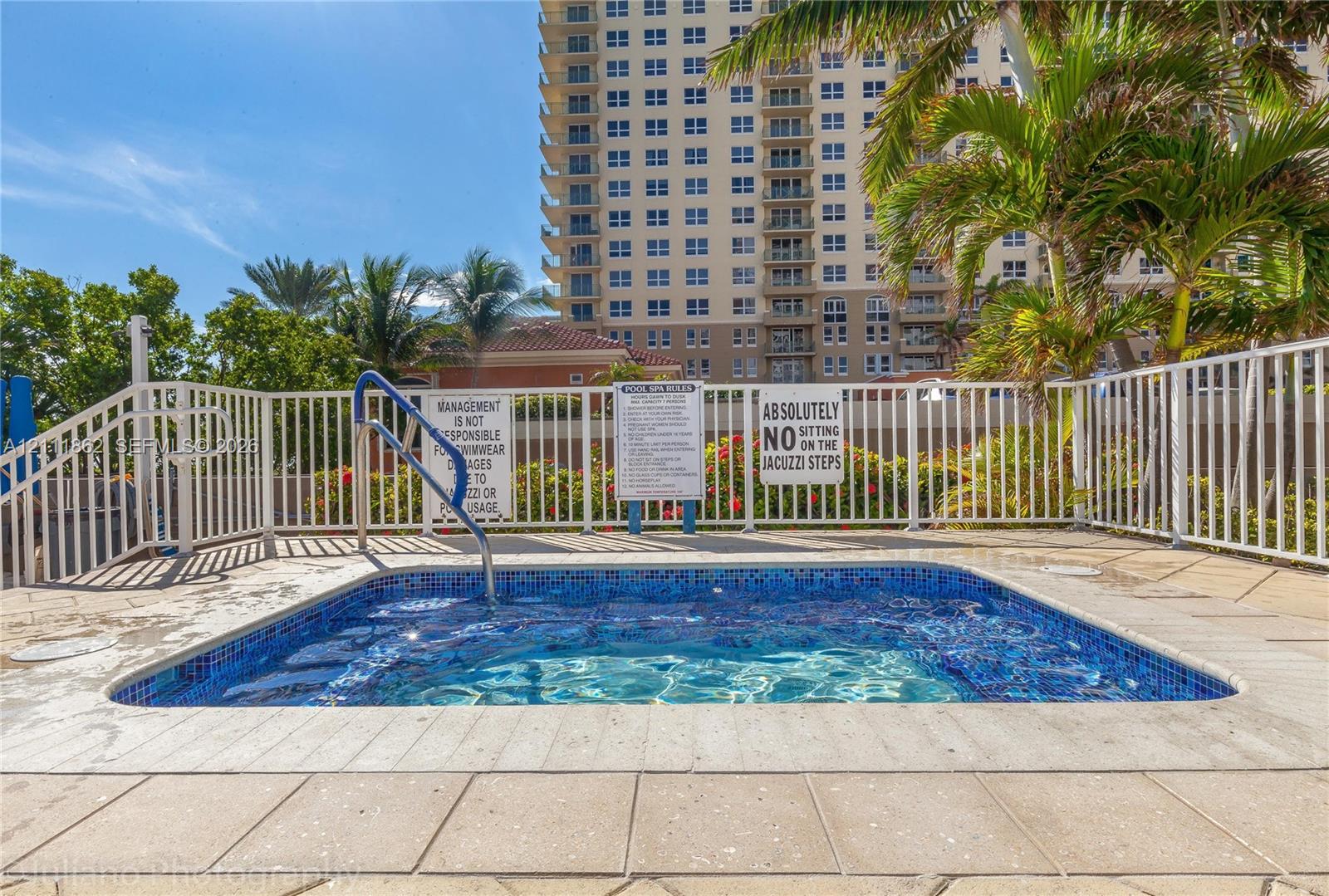 In-ground spa with blue mosaic tile finish, metal handrail, and surrounding paver deck enclosed by white metal fencing