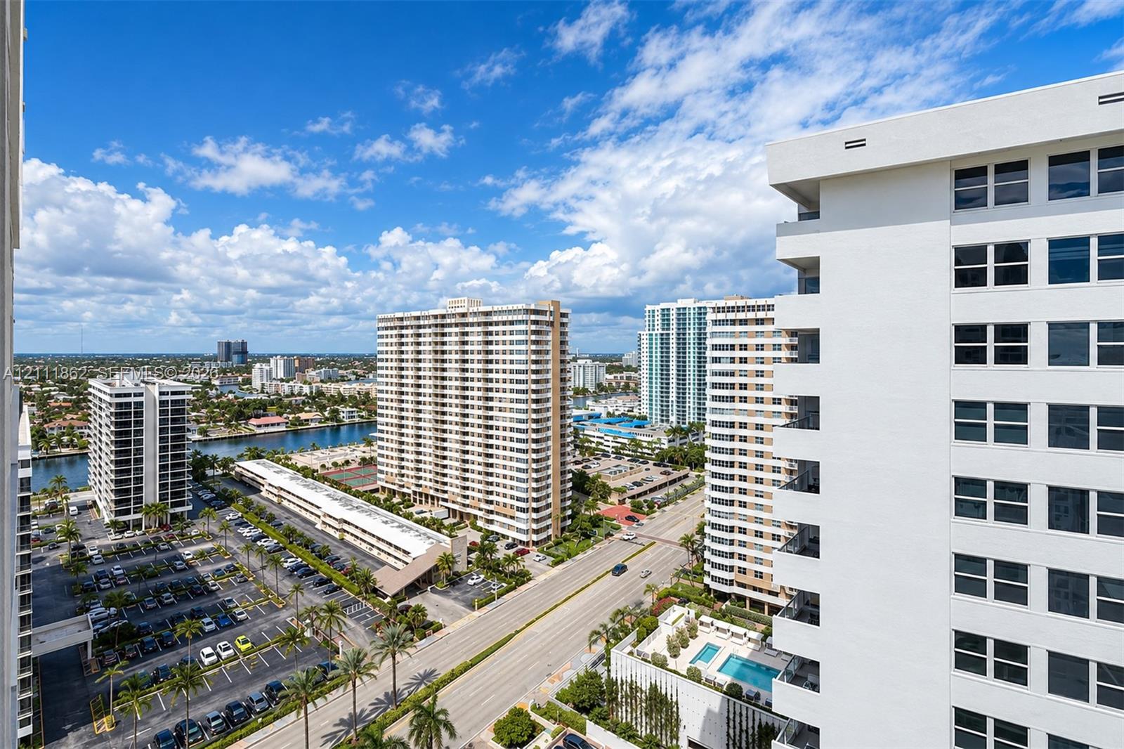 Elevated exterior perspective featuring high-rise concrete architecture, palm tree landscaping, paved parking areas, and a community swimming pool