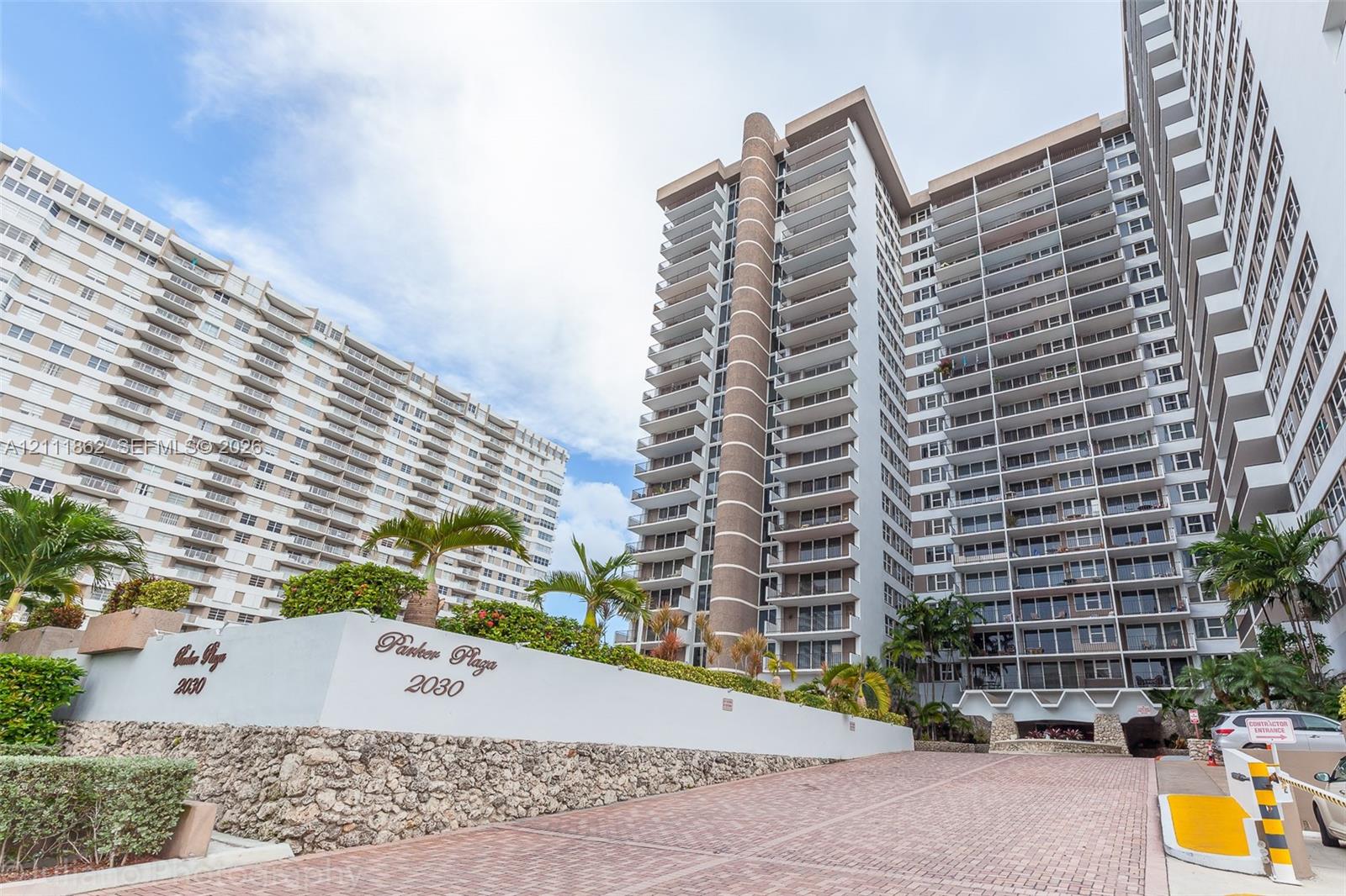 High-rise exterior featuring multi-level private balconies, masonry facade detailing, natural stone-faced retaining walls, paved motor court, and tropical landscaping