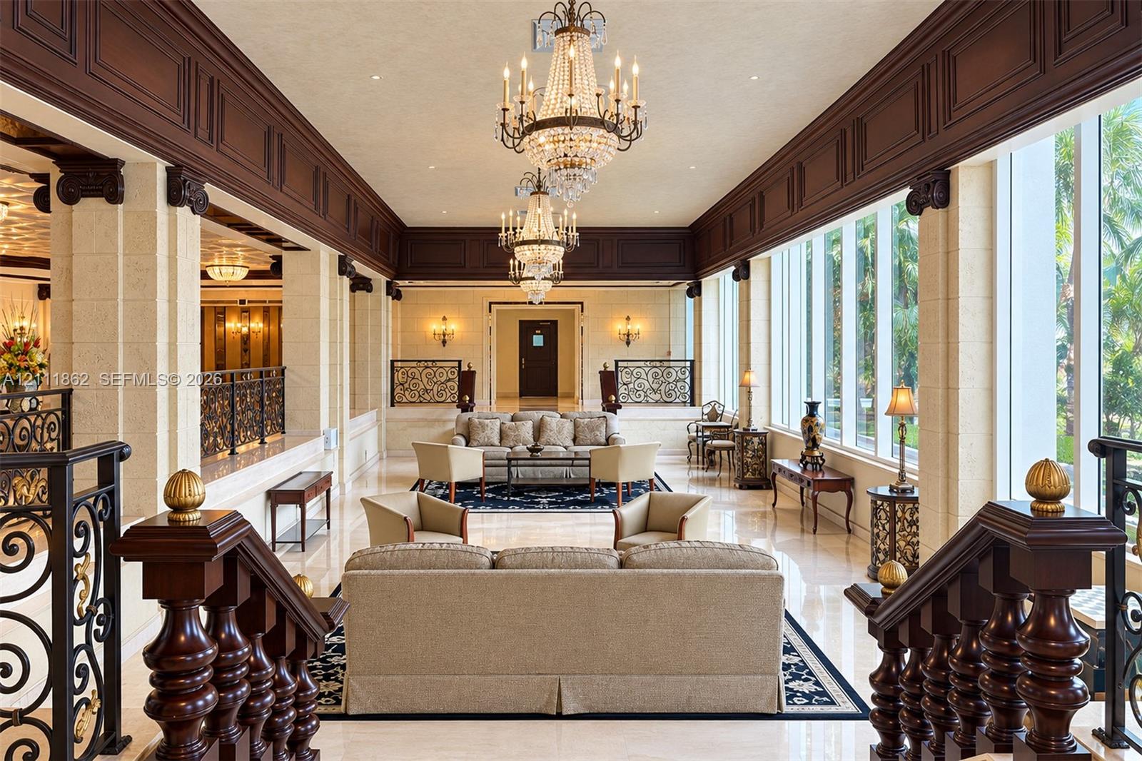Grand interior space featuring high ceilings with custom wood crown molding, dual crystal chandeliers, polished stone flooring, and expansive floor-to-ceiling windows with stone column supports